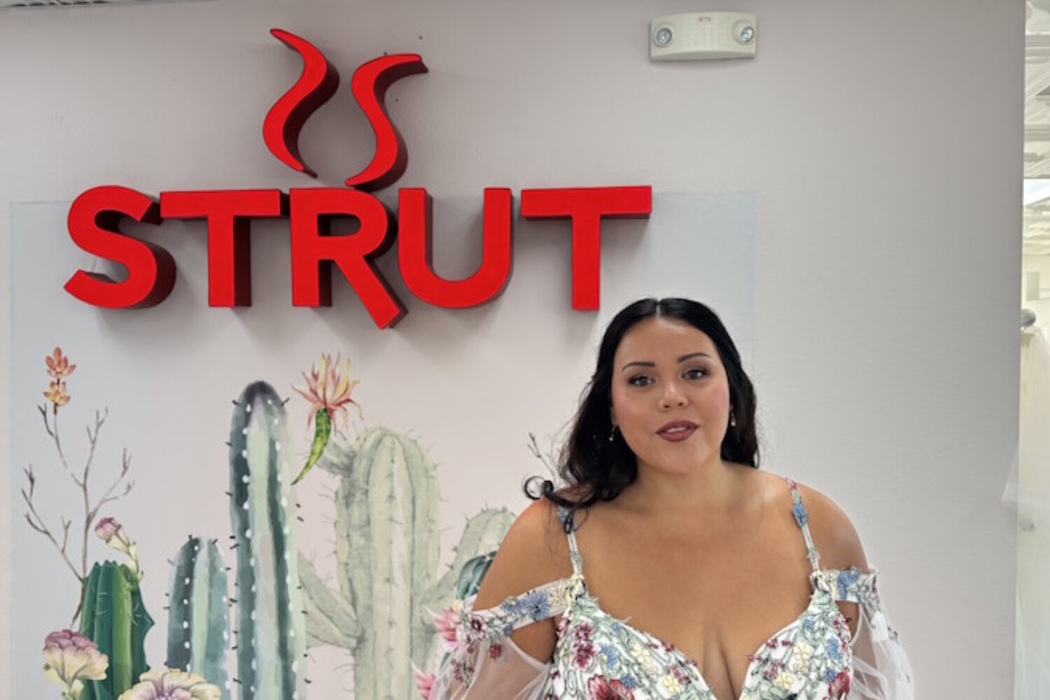 A bride-to-be smiling in front of the 'Strut' logo at Strut Bridal Salon in Chandler, AZ, wearing a floral embroidered wedding dress with off-the-shoulder sleeves and a cactus mural background.