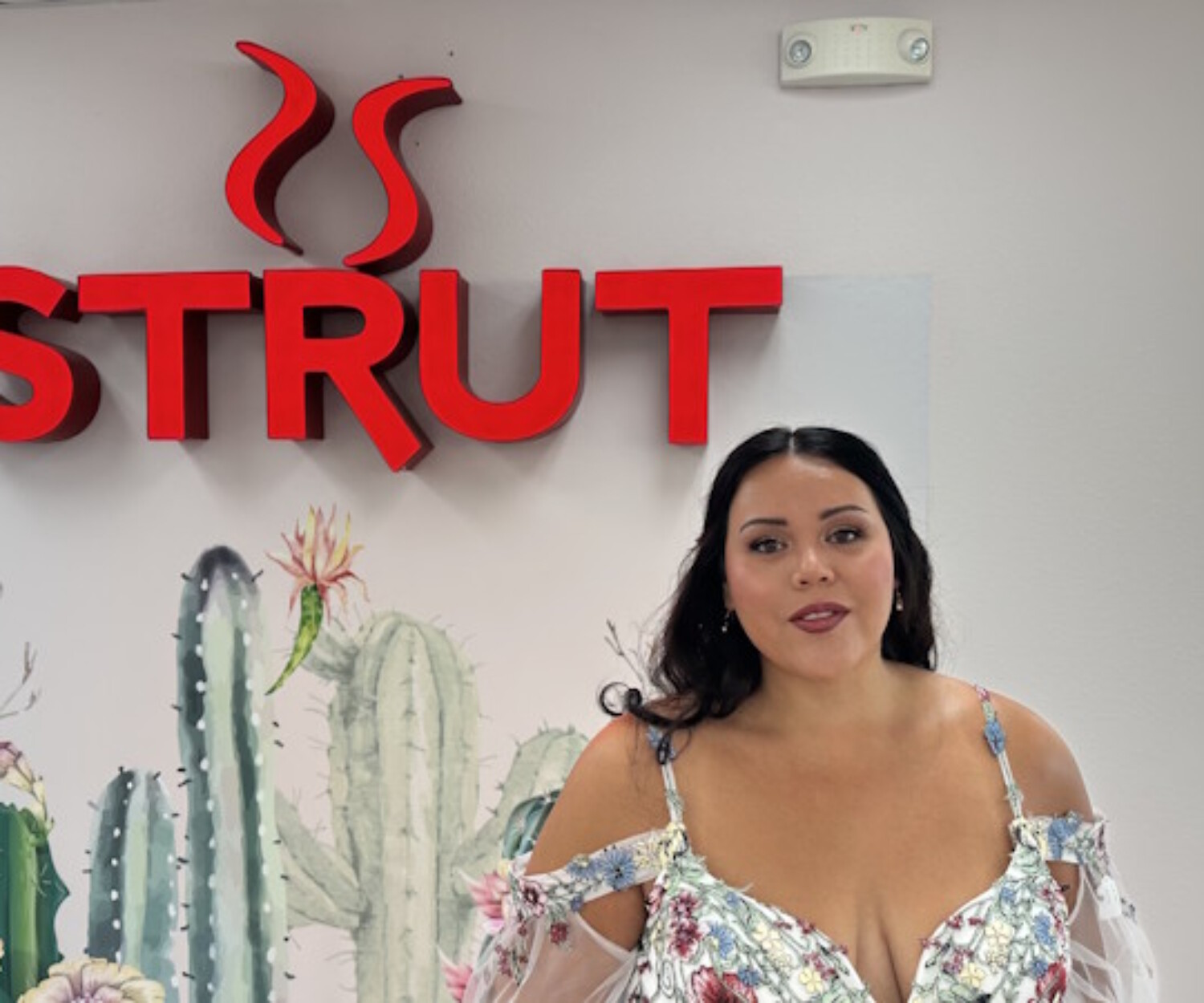 A bride-to-be smiling in front of the 'Strut' logo at Strut Bridal Salon in Chandler, AZ, wearing a floral embroidered wedding dress with off-the-shoulder sleeves and a cactus mural background.