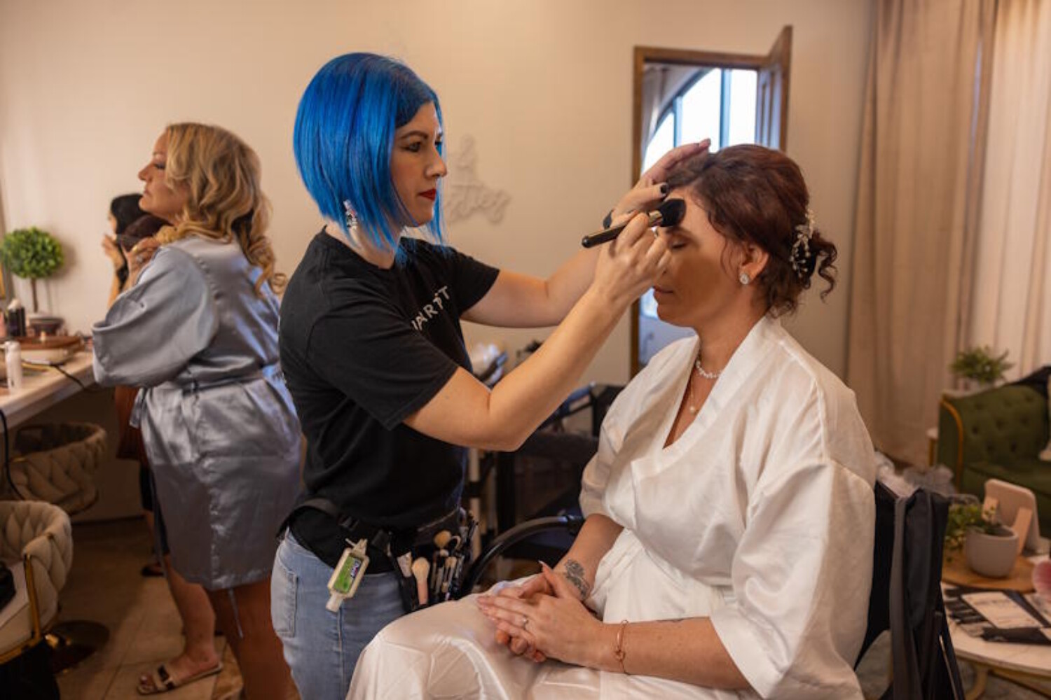 Arizona makeup artist with blue hair applying foundation to a bride in a white robe during a wedding day booking.