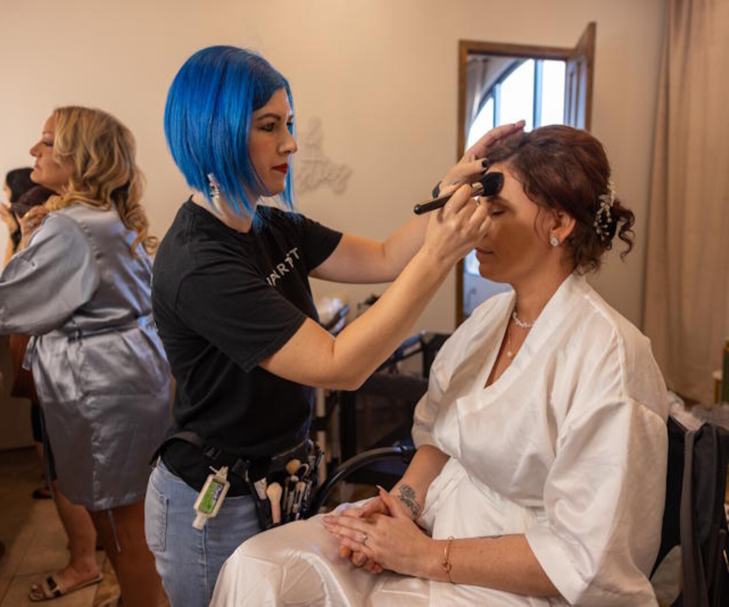 Arizona makeup artist with blue hair applying foundation to a bride in a white robe during a wedding day booking.