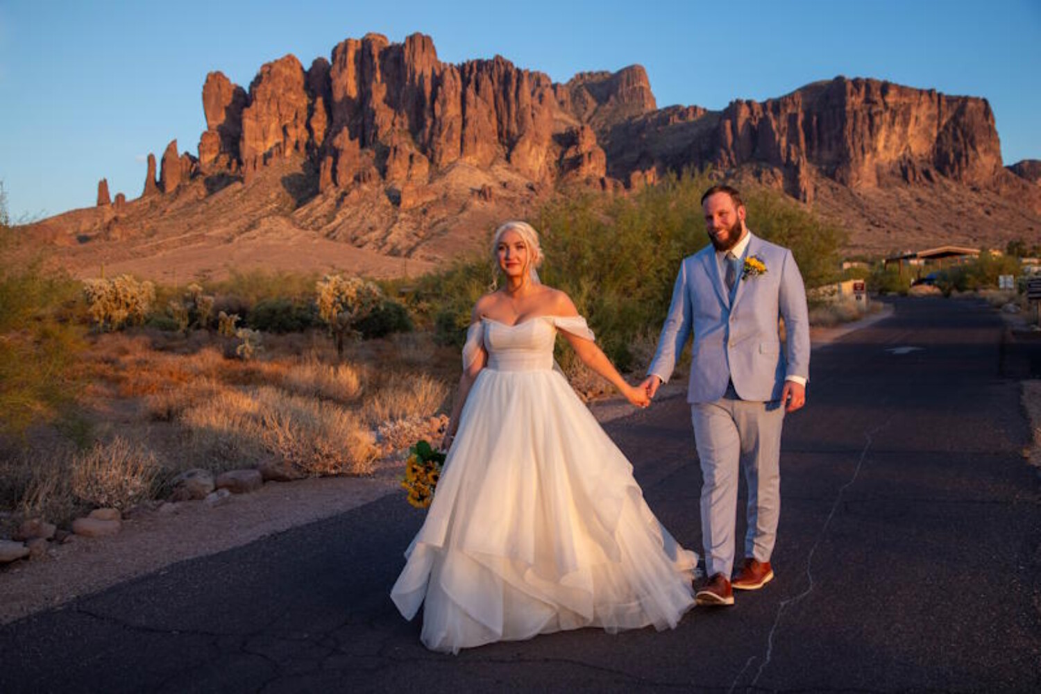 A bride in an off-the-shoulder tulle wedding gown holding hands with a groom in a light blue suit while walking against the backdrop of the rugged Superstition Mountains in Arizona during a golden hour sunset.
