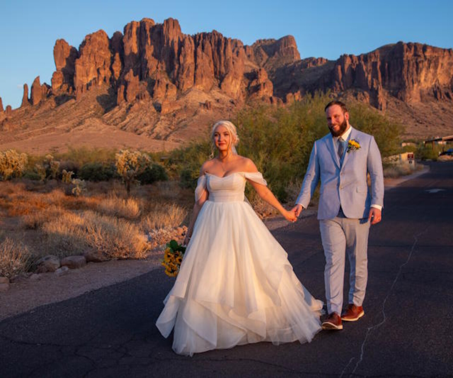 A bride in an off-the-shoulder tulle wedding gown holding hands with a groom in a light blue suit while walking against the backdrop of the rugged Superstition Mountains in Arizona during a golden hour sunset.