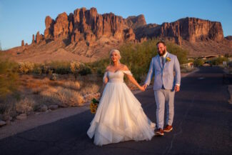 A bride in an off-the-shoulder tulle wedding gown holding hands with a groom in a light blue suit while walking against the backdrop of the rugged Superstition Mountains in Arizona during a golden hour sunset.