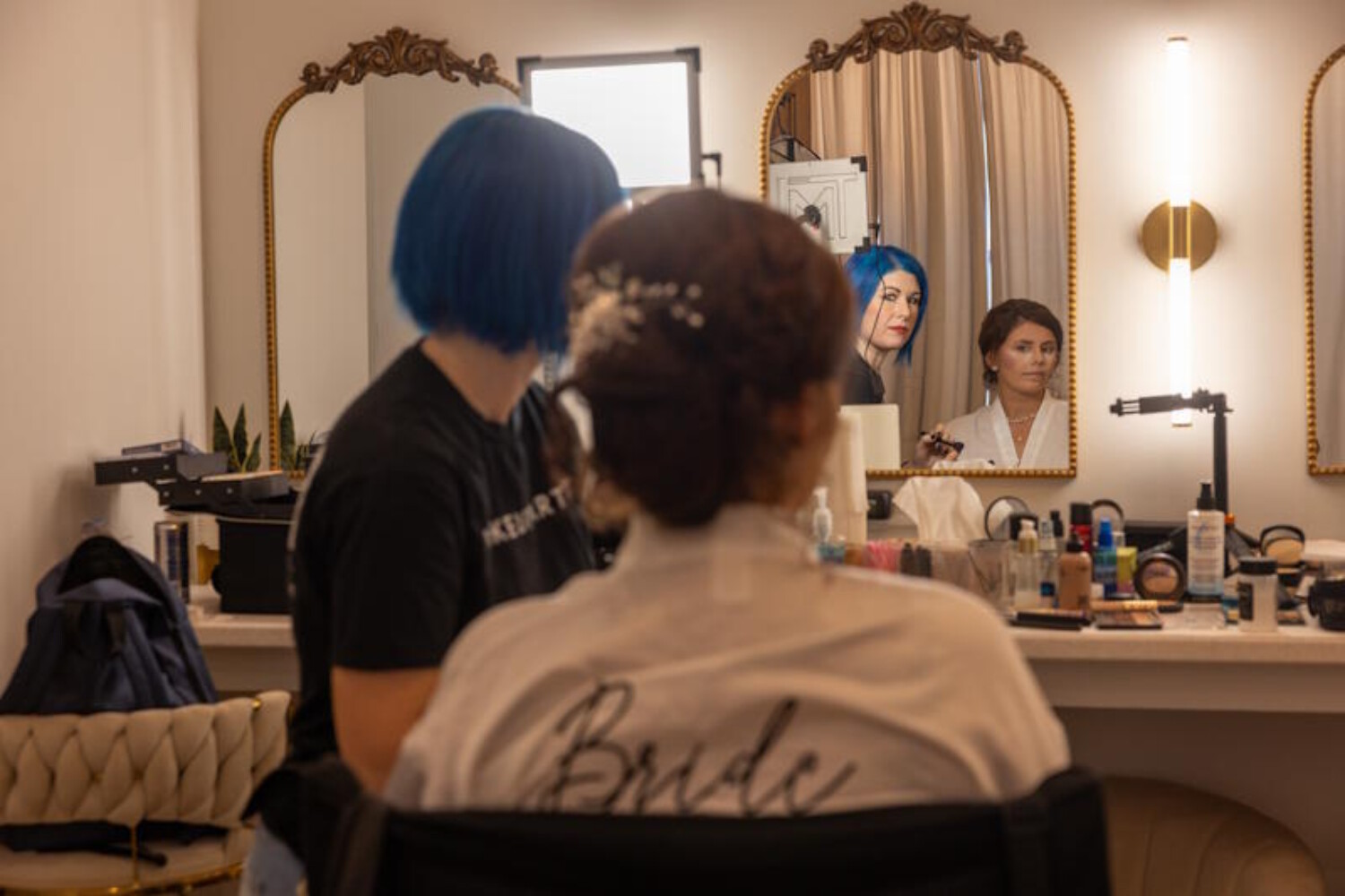 Bride sitting in a bright vanity room while a stylist finishes her elegant updo with hairpins. The bride wears a white robe with “Bride” on the back, surrounded by mirrors, lights, and beauty products. A calm, behind‑the‑scenes moment of wedding preparation captured in warm detail.