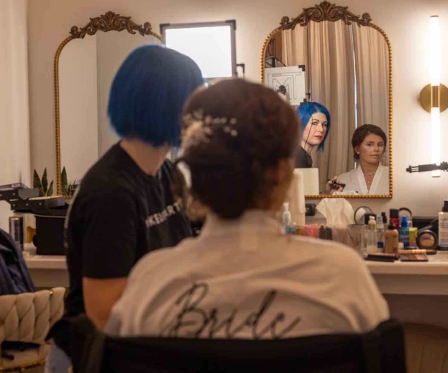Bride sitting in a bright vanity room while a stylist finishes her elegant updo with hairpins. The bride wears a white robe with “Bride” on the back, surrounded by mirrors, lights, and beauty products. A calm, behind‑the‑scenes moment of wedding preparation captured in warm detail.