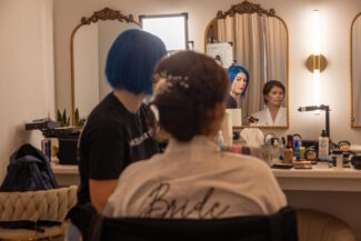 Bride sitting in a bright vanity room while a stylist finishes her elegant updo with hairpins. The bride wears a white robe with “Bride” on the back, surrounded by mirrors, lights, and beauty products. A calm, behind‑the‑scenes moment of wedding preparation captured in warm detail.