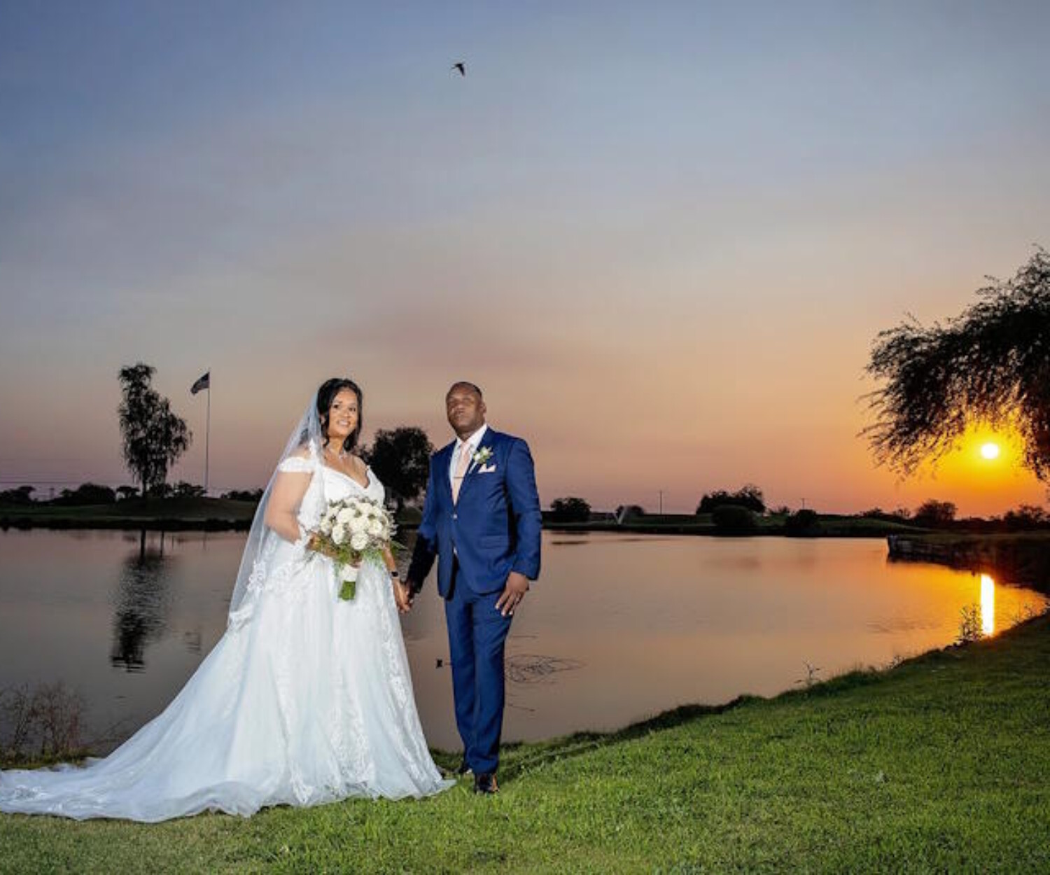 ride in a white lace gown and long veil holding a white bouquet, standing with her groom in a navy suit beside a lake at sunset in Arizona