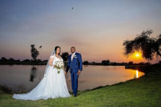 ride in a white lace gown and long veil holding a white bouquet, standing with her groom in a navy suit beside a lake at sunset in Arizona